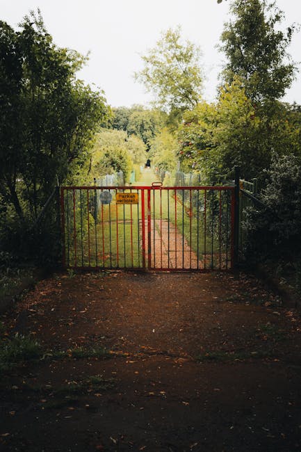 A red metal barrier gate blocks a narrow pathway leading into a shared access garden, with a yellow warning sign attached. The pathway is flanked by dense, mature trees and lush greenery, including ta