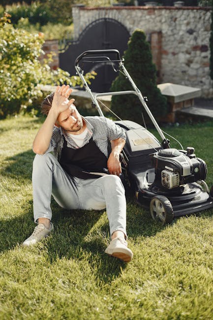 A young man sitting on a well-maintained grassy lawn, appearing tired with one hand on his forehead, beside a black petrol-powered lawnmower. The garden features a stone wall with an arched gate in th